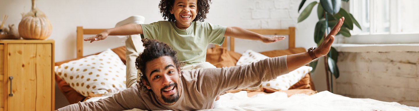 A smiling Dad and his son make airplane motions while laying on a clean comforter.