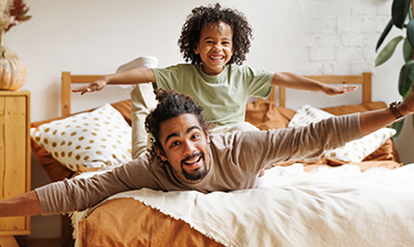 A smiling Dad and his son make airplane motions while laying on a clean comforter.