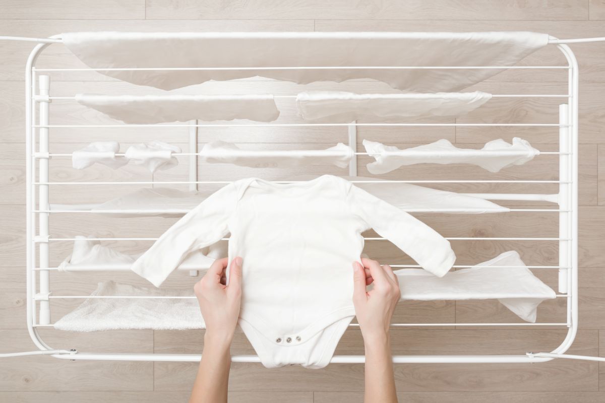 A person laying out a white-colored long-sleeve baby onesie on top of a laundry drying rack after washing. Other white clothing items, including baby clothes, are already hanging on the drying rack.