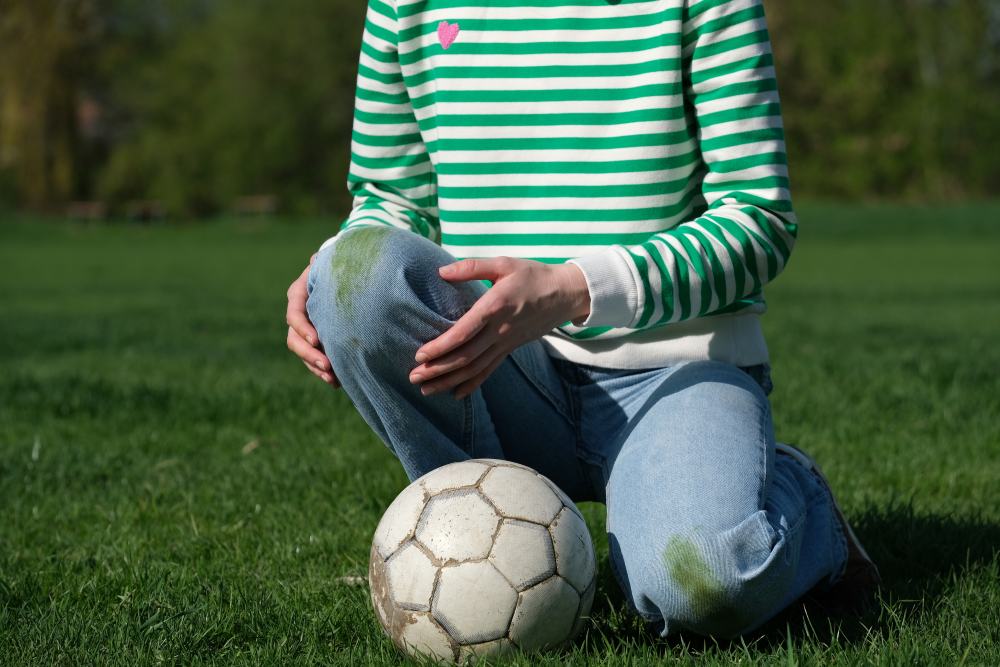 Person kneeling on a grass soccer field with a soccer ball and visible grass stains on clothing.